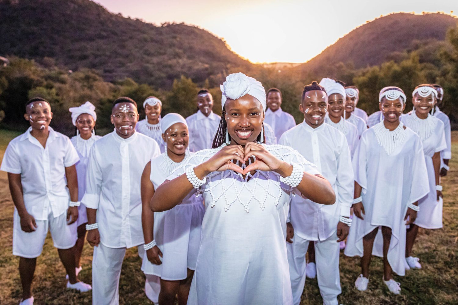 Die Mzansi Youth Choir staan in &rsquo;n veld met sononder, almal in wit, met die sangeres voor wat &rsquo;n hartjie met haar hande maak